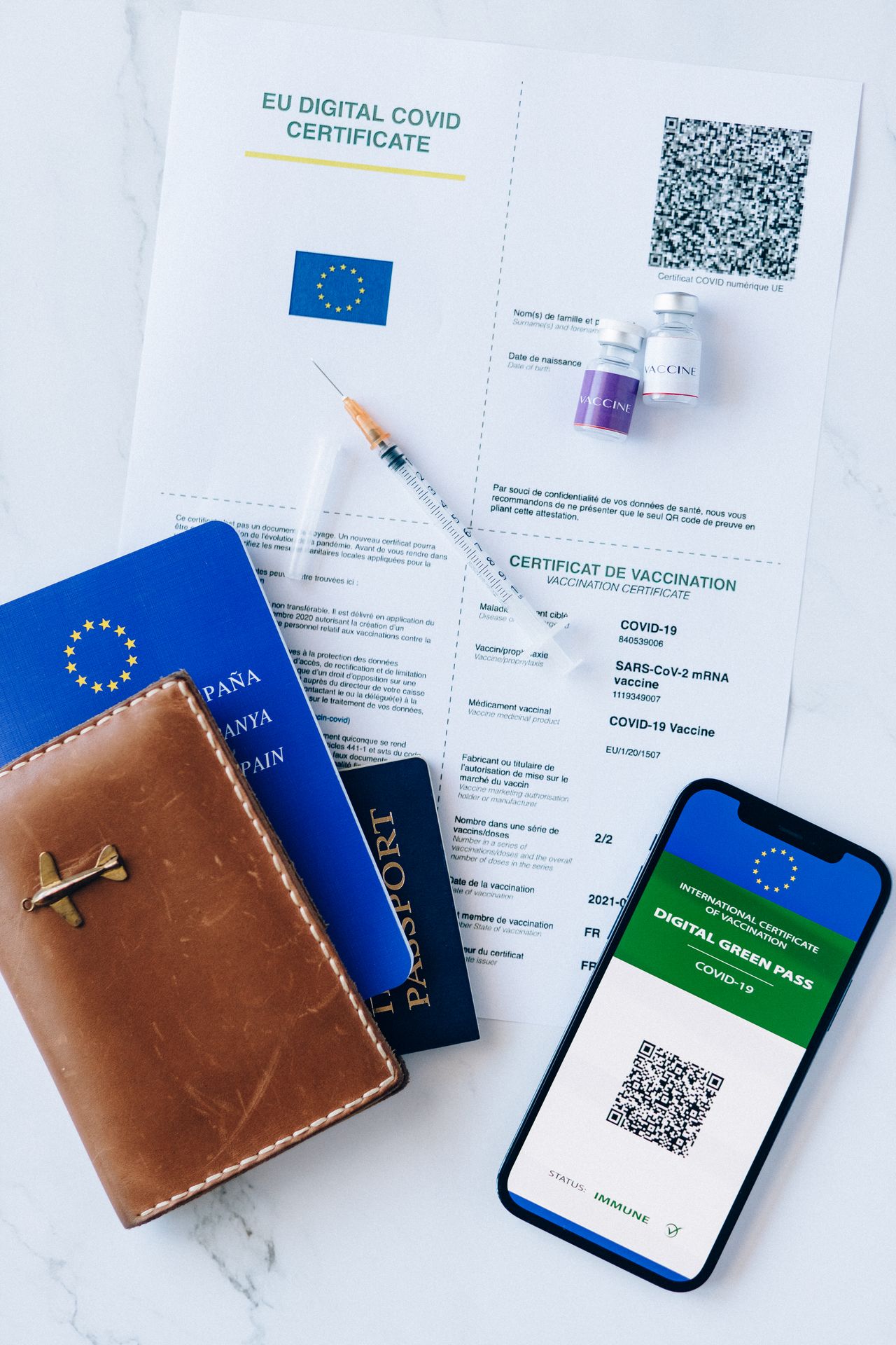 EU digital COVID vaccination certificate on a marble surface with two passports, a leather passport wallet, syringe, vaccine vials and a phone showing a digital green pass