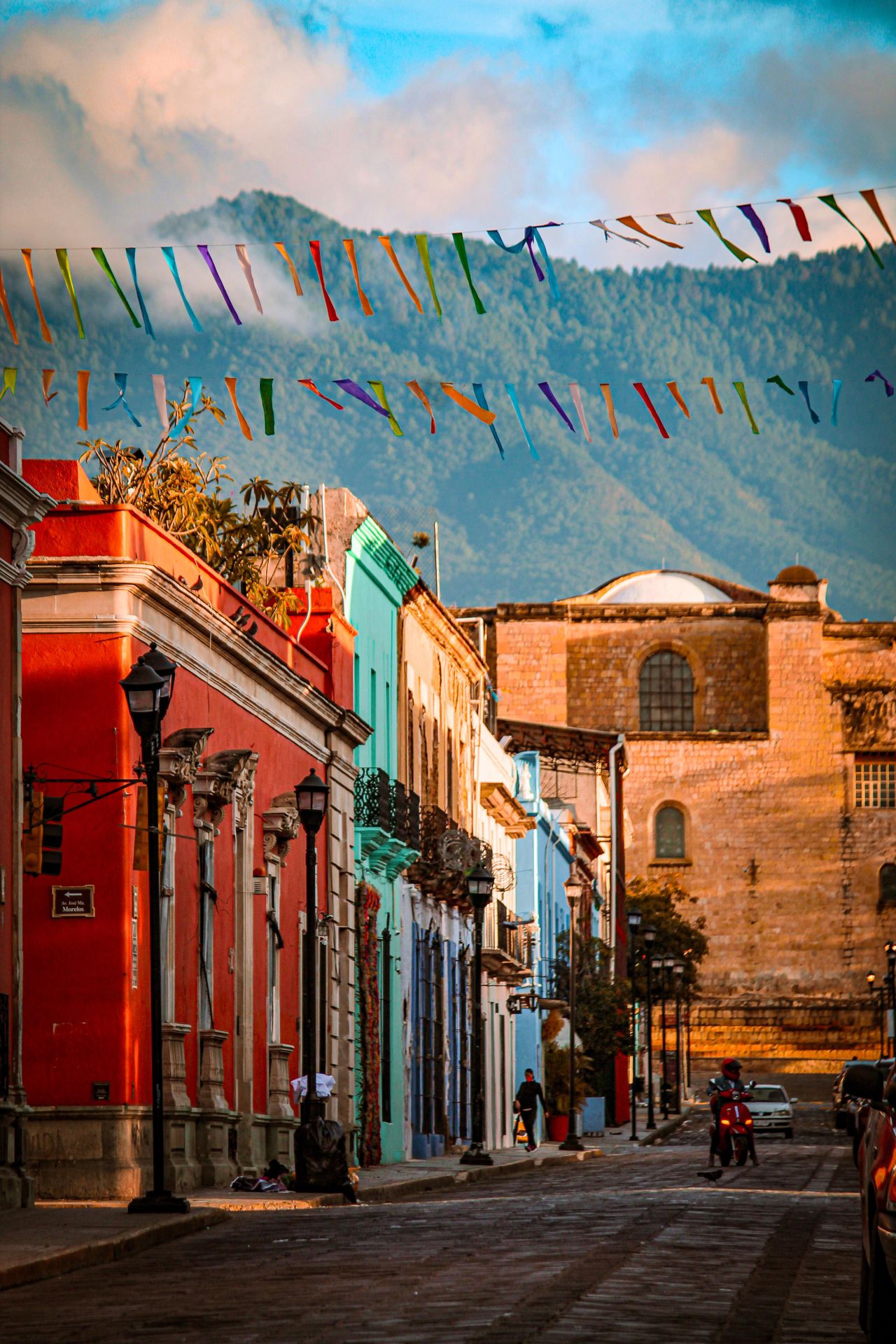 Colourful papel picado bunting strung above a cobblestone street lined with red and turquoise colonial buildings in Oaxaca, Mexico, with misty mountains behind