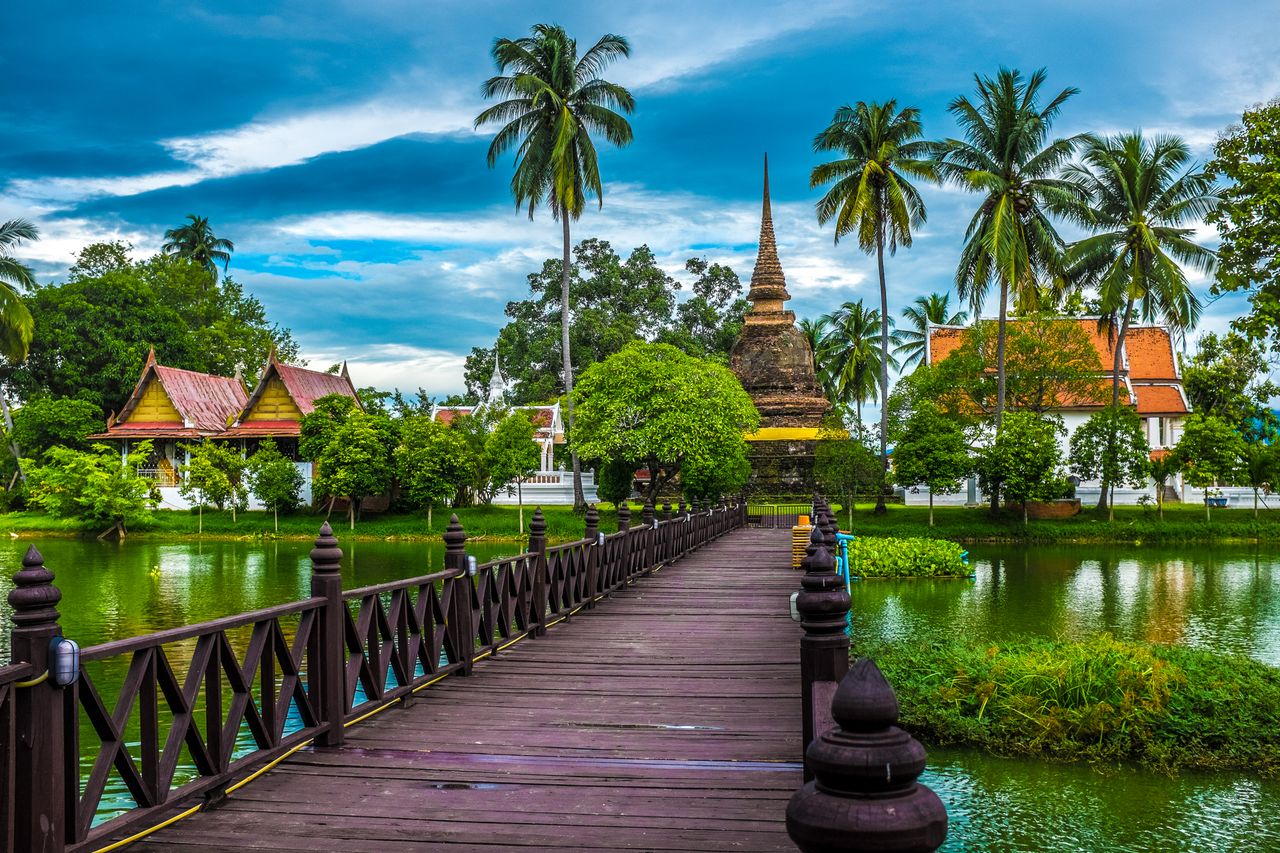 Wooden footbridge leading to a stupa surrounded by palm trees and temple pavilions in Sukhothai, Thailand