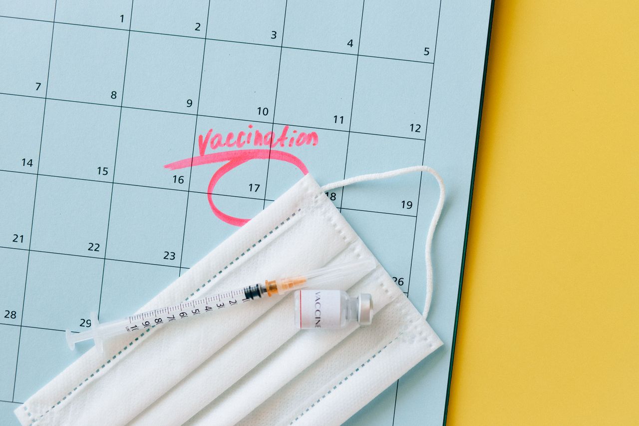 Overhead view of a calendar with the word 'vaccination' handwritten and circled in pink marker, alongside a medical face mask, vaccine vial and syringe