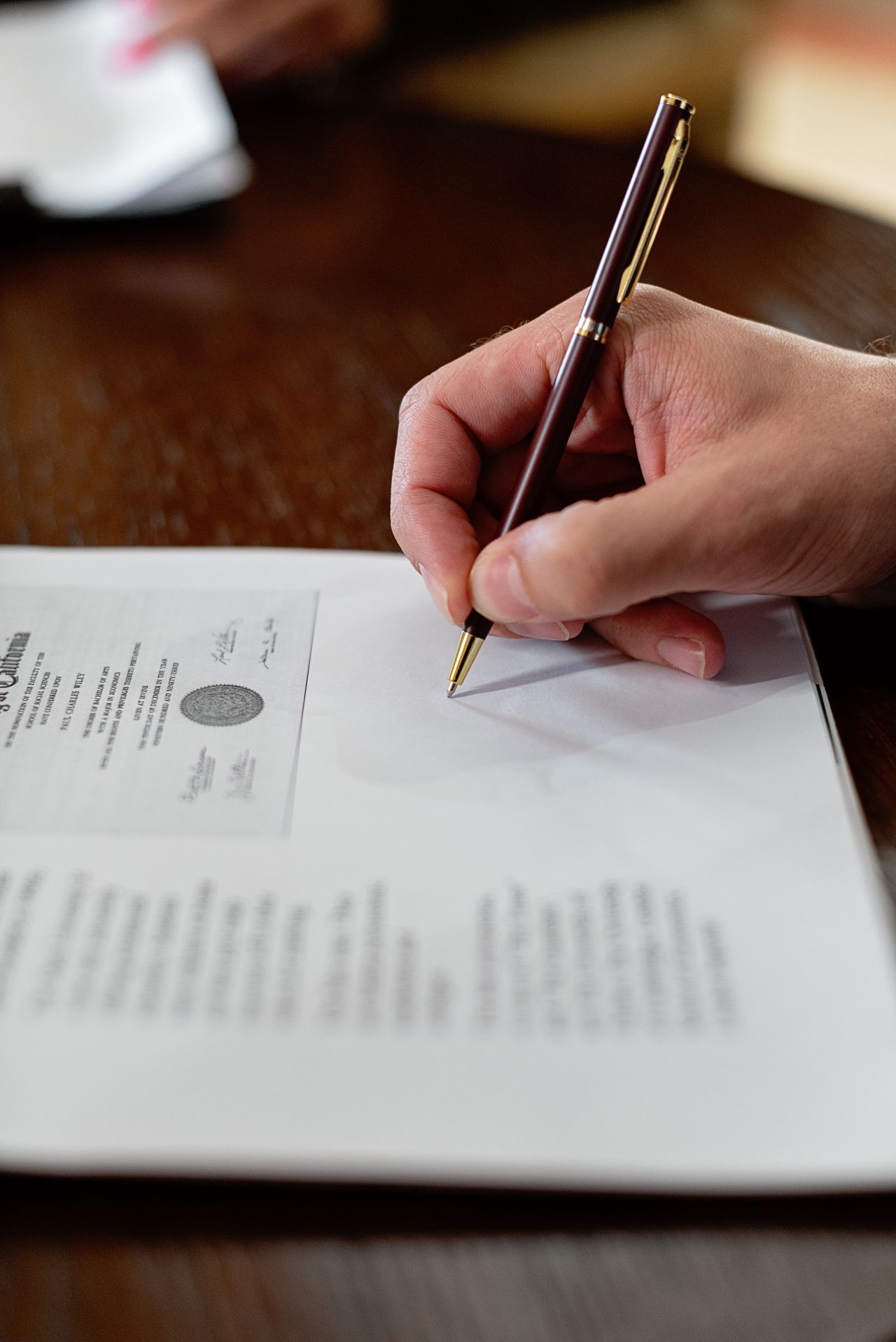 Close-up of a hand signing an official certificate document with a dark red and gold pen at a wooden desk